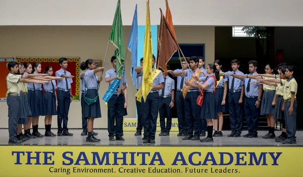 The Samhita Academy The Samhita Academy, Bangalore — pupils in uniform holding house flags on a stage above the school's name board